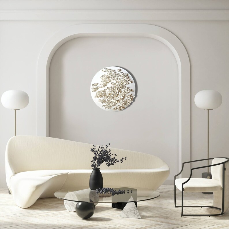 May include: A white, round wall sculpture with a textured, organic design. The sculpture is hanging on a white wall in a modern living room with a white sofa, a coffee table, and two floor lamps.