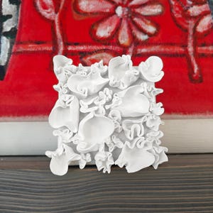 May include: A square, white, textured art piece with a three-dimensional floral design. The artwork is set against a backdrop of a red painting with white floral patterns. The piece is displayed on a dark wooden surface.