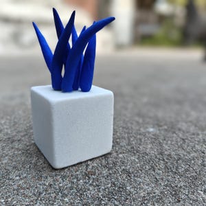 May include: A small white cube planter with a cluster of blue ceramic spikes. The planter is sitting on a gray concrete surface.