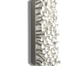 Puede incluir: Escultura mural rectangular blanca con superficie texturizada. La escultura presenta numerosas formas pequeñas, redondeadas, con forma de pétalo que sobresalen de una base rectangular. La pieza está montada en una pared blanca, creando una sombra.