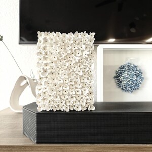 May include: A rectangular white floral sculpture composed of numerous small, textured flowers. The sculpture sits atop a black, textured rectangular box, likely a speaker or soundbar, on a wooden surface. A framed blue floral artwork is in the background.