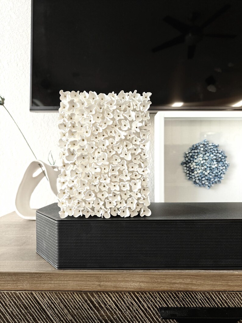 May include: A rectangular white floral sculpture composed of numerous small, textured flowers. The sculpture sits atop a black, textured rectangular box, likely a speaker or soundbar, on a wooden surface. A framed blue floral artwork is in the background.