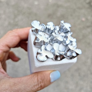 May include: A white cube with a silver metal design on top. The design is made up of multiple small, flower-shaped pieces.