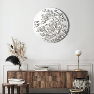 May include: A white wall with a large white circular wall sculpture. The sculpture is made of many small white shapes that resemble flowers. A wooden dresser with three drawers is in the foreground. The dresser is made of dark wood with brass handles. A vase with dried flowers is on the dresser. A lamp with a white globe is on the right side of the dresser.
