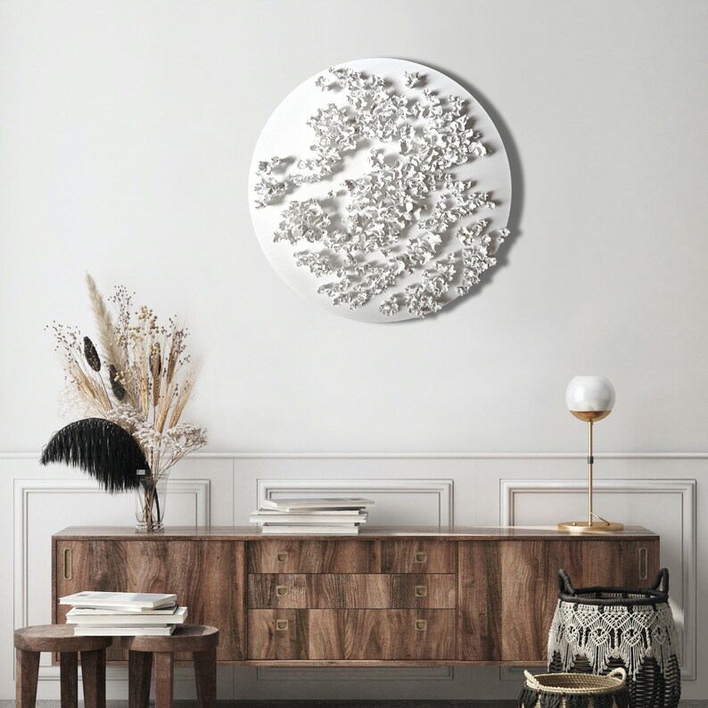 May include: A white wall with a large white circular wall sculpture. The sculpture is made of many small white shapes that resemble flowers. A wooden dresser with three drawers is in the foreground. The dresser is made of dark wood with brass handles. A vase with dried flowers is on the dresser. A lamp with a white globe is on the right side of the dresser.