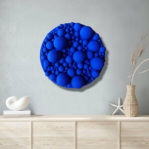May include: A round, textured wall art piece featuring a vibrant blue color. The artwork is composed of numerous spheres of varying sizes, creating a three-dimensional effect. The piece is mounted on a light gray wall, adding a modern touch to the decor.