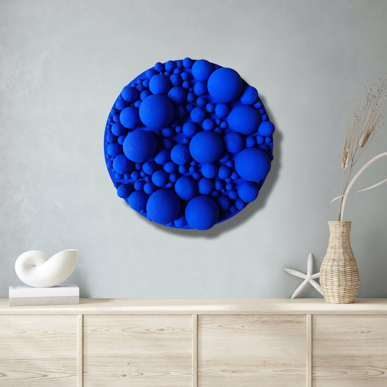 May include: A round, textured wall art piece featuring a vibrant blue color. The artwork is composed of numerous spheres of varying sizes, creating a three-dimensional effect. The piece is mounted on a light gray wall, adding a modern touch to the decor.