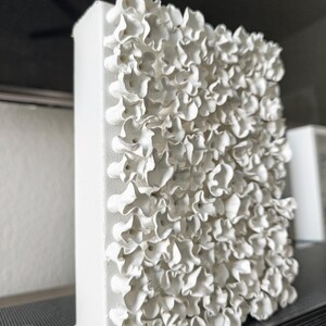 May include: A white, textured wall art piece with a three-dimensional floral design. The artwork is rectangular and appears to be made of a fabric-like material. The flowers are clustered together, creating a layered effect.