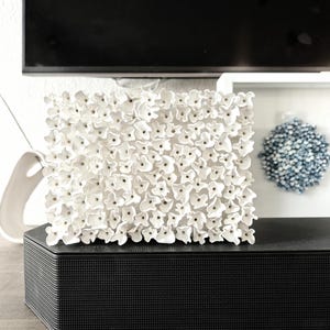 May include: A square, white floral wall art piece. The artwork is composed of numerous small, three-dimensional white flowers. It sits atop a black, textured speaker on a wooden surface. A blue decorative sphere is visible in the background.
