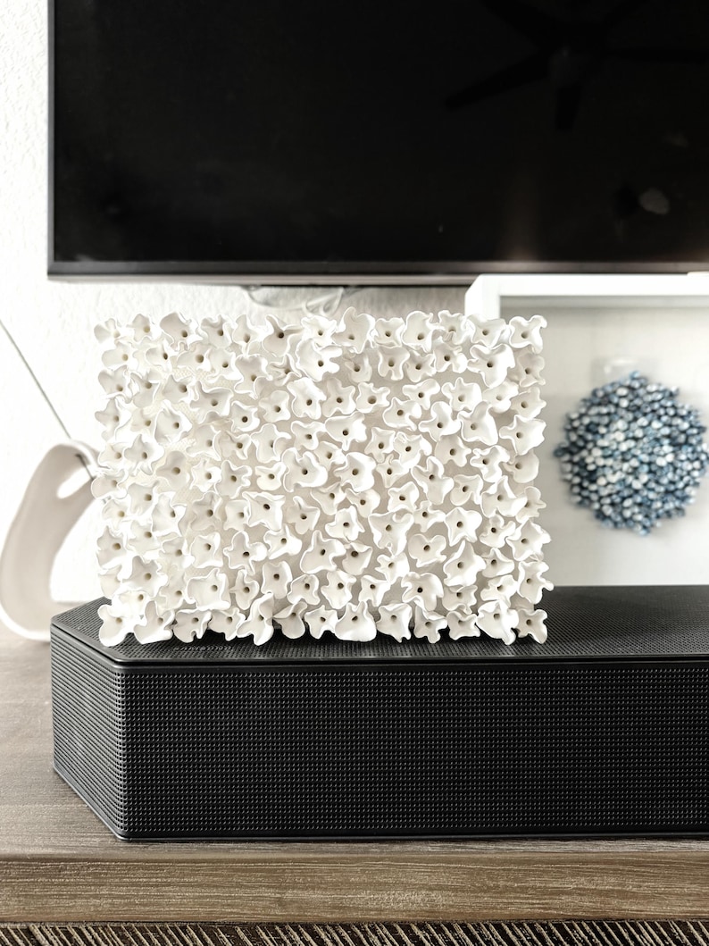 May include: A square, white floral wall art piece. The artwork is composed of numerous small, three-dimensional white flowers. It sits atop a black, textured speaker on a wooden surface. A blue decorative sphere is visible in the background.