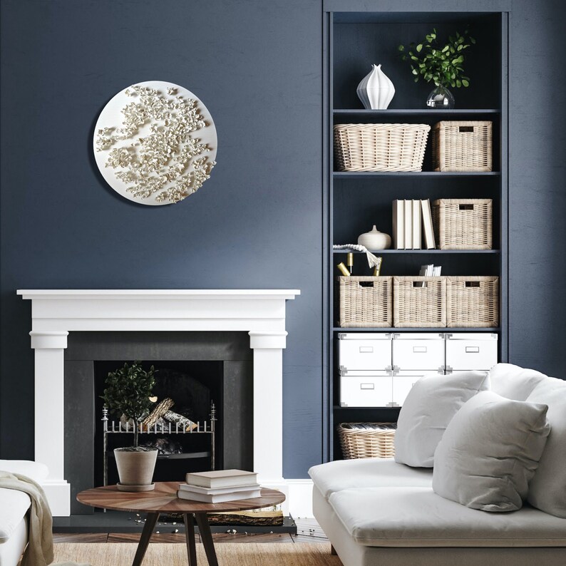 May include: A white circular wall sculpture with a textured, organic design hangs above a white fireplace in a living room with a white couch and a wooden coffee table. The room has dark blue walls and a built-in bookshelf with wicker baskets and books.