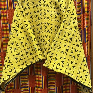 May include: A yellow poncho-style top with a black geometric pattern. The garment has a wide neckline and a black trim along the edges. The background features a colorful patterned fabric.