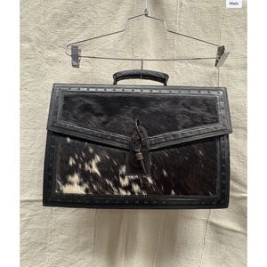 以下が含まれることがあります： 黒と白の牛革のブリーフケース。革製のハンドルとトリムが付いています。ブリーフケースには金属製の留め金が付いており、金属製のハンガーから吊り下げられています。