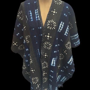 Puede incluir: Chaqueta estilo kimono con estampado azul oscuro y negro con diseños blancos de estrellas y geométricos. La chaqueta tiene cuello en V y flecos en el borde inferior. La prenda se exhibe en un maniquí sobre un fondo negro.
