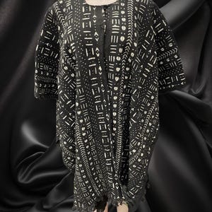 May include: Black and white patterned tunic with short sleeves. The tunic features a geometric design with white shapes on a black background. The garment has a fringed hem and is displayed on a mannequin.