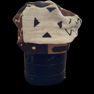 May include: A knitted beanie hat with a geometric pattern in white, brown, and black. The hat features a folded brim and a textured design, perfect for cold weather. The hat is displayed on a dark blue cylindrical object against a black background.