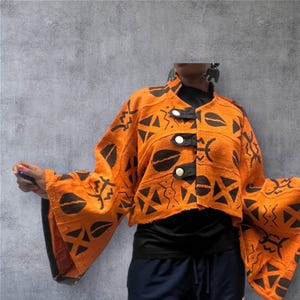 Puede incluir: Chaqueta corta naranja con patrones geométricos negros y mangas anchas. La chaqueta presenta un cuello mandarín, ribetes negros y tres botones blancos. La prenda está hecha de una tela texturizada.