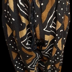 Puede incluir: Pantalones negros con un patrón repetido de formas marrones y blancas. Los pantalones tienen cintura con cordón y se exhiben en un maniquí. La tela parece ser un material tejido.