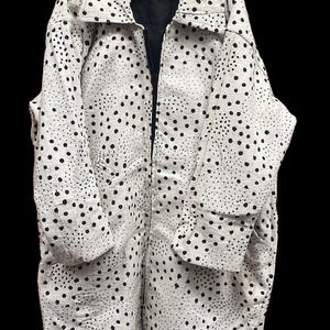 Puede incluir: Mono blanco con estampado de lunares negros. El mono tiene cuello, mangas largas y cremallera completa. La prenda está colgada de una percha negra sobre un fondo negro.