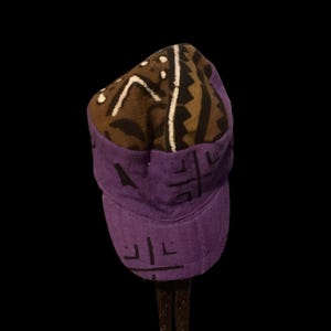 Puede incluir: Gorra morada y marrón estampada con visera corta. La gorra presenta un diseño geométrico en marrón, negro y blanco en la parte superior, y un color morado liso en la visera y la parte inferior.