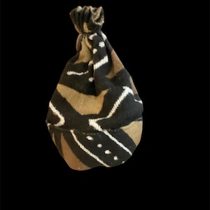 Op de afbeelding: Een kleine, bedrukte muts in bruin-, zwart- en wittinten. De muts heeft een geknoopte bovenkant en een herhalend geometrisch ontwerp. De stof lijkt zacht en getextureerd, geschikt voor een baby of een klein kind.