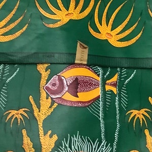 Op de afbeelding: Groene stof met een herhaald patroon van gele koraalachtige vormen en witte takachtige lijnen. Een visvormige applicatie met bruine en gele details is gecentreerd op de stof. Het thema is marien.
