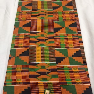 Pode incluir: Um tecido colorido com um padrão geométrico em tons de laranja, verde, preto e marrom. O tecido tem uma textura tecida e está dobrado em uma pilha. O tecido tem uma etiqueta que diz "AFRICA".