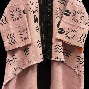 May include: A pink and black patterned jacket with a unique design. The jacket features a light pink fabric with black geometric and sun-like patterns. The front has a black panel with button closures. The sleeves are three-quarter length.