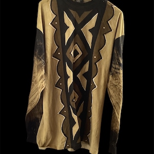 Könnte beinhalten: Beigefarbenes Langarmshirt mit einem geometrischen Muster in Schwarz und Braun. Das Shirt hat einen hohen Ausschnitt und ein einzigartiges Muster auf der Vorderseite. Die Ärmel haben ein dunkles, verblasstes Design. Auf einem Holzkleiderbügel.