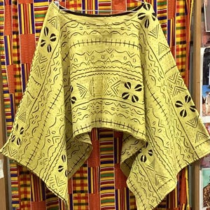 May include: A light yellow poncho-style top with black geometric patterns and floral accents. The top is hanging on a hanger against a colorful patterned fabric background. The design features various shapes and lines.