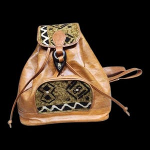 May include: A tan leather backpack with a flap closure and drawstring. The flap and front pocket feature patterned fabric in shades of green, black, and white. The bag has a dark toggle closure and adjustable straps.