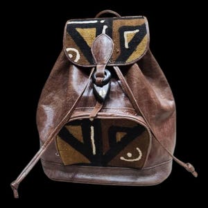 May include: Brown leather backpack with a flap closure and a front pocket. The flap and pocket feature a geometric pattern in black, tan, and white. The backpack has a drawstring closure and adjustable straps.