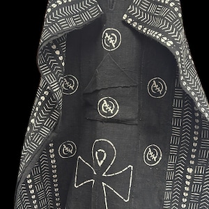 May include: Black tunic with white geometric patterns and an Ankh symbol. The tunic features a V-neck, a pocket, and circular designs. The sleeves and edges are adorned with repeating arrow and line patterns. The garment is displayed on a hanger against a black background.