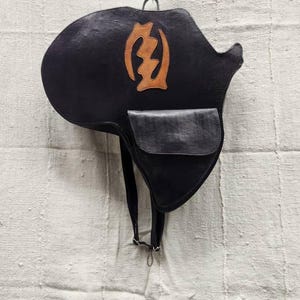 May include: Black leather saddlebag with a brown emblem and a small pocket. The bag is hanging from a metal hook. The saddlebag has a strap with a silver clasp.