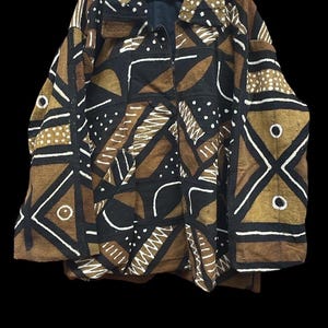 Puede incluir: Chaqueta estampada sobre fondo negro. La chaqueta presenta un diseño geométrico en marrón, negro y blanco. El diseño incluye formas como triángulos, círculos y líneas. La chaqueta está colgada de una percha negra.