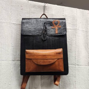 May include: Black leather backpack with a brown leather flap pocket and straps. The flap features a brown ankh symbol and a leather closure. The bag is hanging against a textured white background.