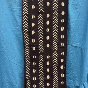 Könnte beinhalten: Schwarzer Schal mit einem weißen geometrischen Muster aus Pfeilen und ovalen Formen. Der Schal hat einen Fransenrand und hängt vor einem blauen Hintergrund.