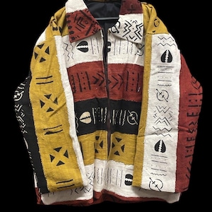 Puede incluir: Chaqueta colorida con cremallera negra, con un diseño de patchwork en amarillo, rojo, negro y blanco. La chaqueta está adornada con varios patrones geométricos y simbólicos, creando un aspecto único y artístico. Fondo negro.
