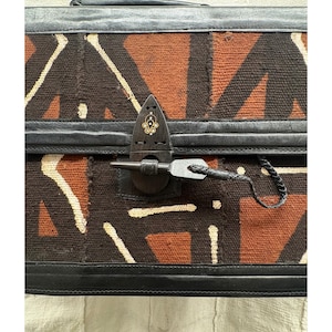 May include: A rectangular, patterned bag with a geometric design in brown, orange, and white. The bag features black leather trim and a decorative metal clasp. The bag has a handle and a key attached to the clasp.