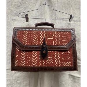 May include: A rectangular briefcase with a brown leather handle and trim. The main body features a red and white patterned fabric. The briefcase is secured with a leather clasp and is suspended from a metal hanger.