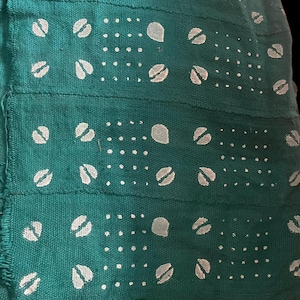 Puede incluir: Primer plano de una tela turquesa con un patrón repetitivo de formas blancas. El diseño incluye pequeños puntos y formas estilizadas más grandes que se asemejan a semillas o frijoles. La tela parece cosida a mano, con costuras visibles y una superficie texturizada.