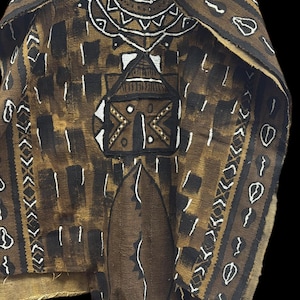 May include: A brown and black patterned tunic with white geometric designs. The tunic features a V-neck and a central rectangular design with a house-like motif. The fabric has a textured appearance, and the overall style is reminiscent of traditional African attire.