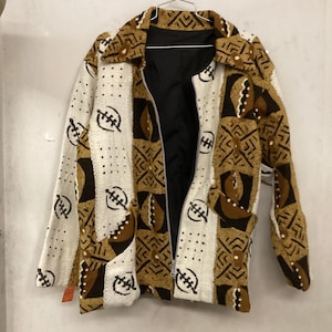 May include: A long-sleeve jacket featuring a textured pattern in shades of brown, cream, and black. The garment has a collared neckline, a full-length front zipper, and a solid black interior lining. The fabric displays repeating geometric shapes.