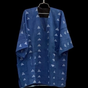 May include: A blue tunic with a V-neckline and short sleeves, featuring a repeating pattern of light blue triangles across the front. The garment includes two rectangular patch pockets on the lower front and is displayed on a metal hanger.