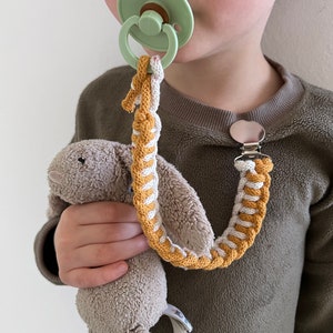 May include: A child wearing a brown sweater with a dummy attached to a braided cord with a plush toy attached to the other end. The cord is braided with yellow and white yarn.