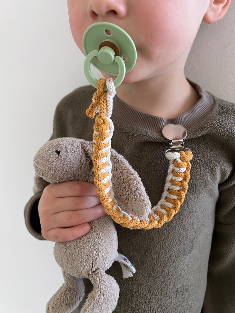 May include: A child wearing a brown sweater with a dummy attached to a braided cord with a plush toy attached to the other end. The cord is braided with yellow and white yarn.