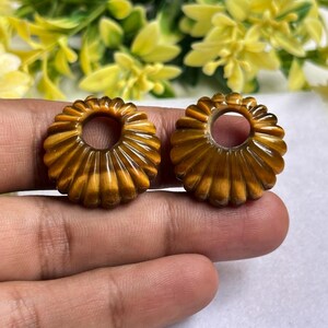 May include: Two circular, golden-brown tiger's eye beads featuring a carved, ribbed, sunburst-like pattern radiating from a central circular hole. These polished, chatoyant gemstone components exhibit natural banding and a glossy, smooth finish.