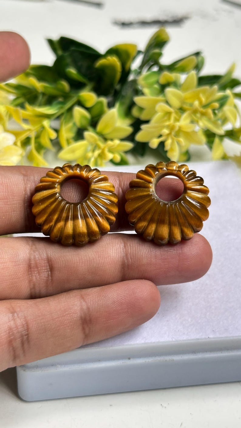 May include: Two circular, golden-brown tiger's eye beads featuring a carved, ribbed, sunburst-like pattern radiating from a central circular hole. These polished, chatoyant gemstone components exhibit natural banding and a glossy, smooth finish.