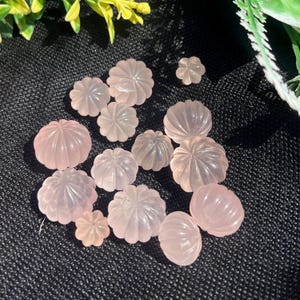 May include: A collection of carved rose quartz gemstones in various sizes and shapes, resembling small pumpkins and flowers. The gemstones are a soft pink color and are arranged on a black textured surface.