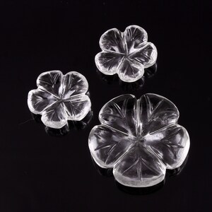 May include: Three clear, flower-shaped crystal carvings. The carvings have five petals with etched details, and vary in size. The largest flower is at the bottom, with two smaller flowers above it. The crystals are on a black surface.